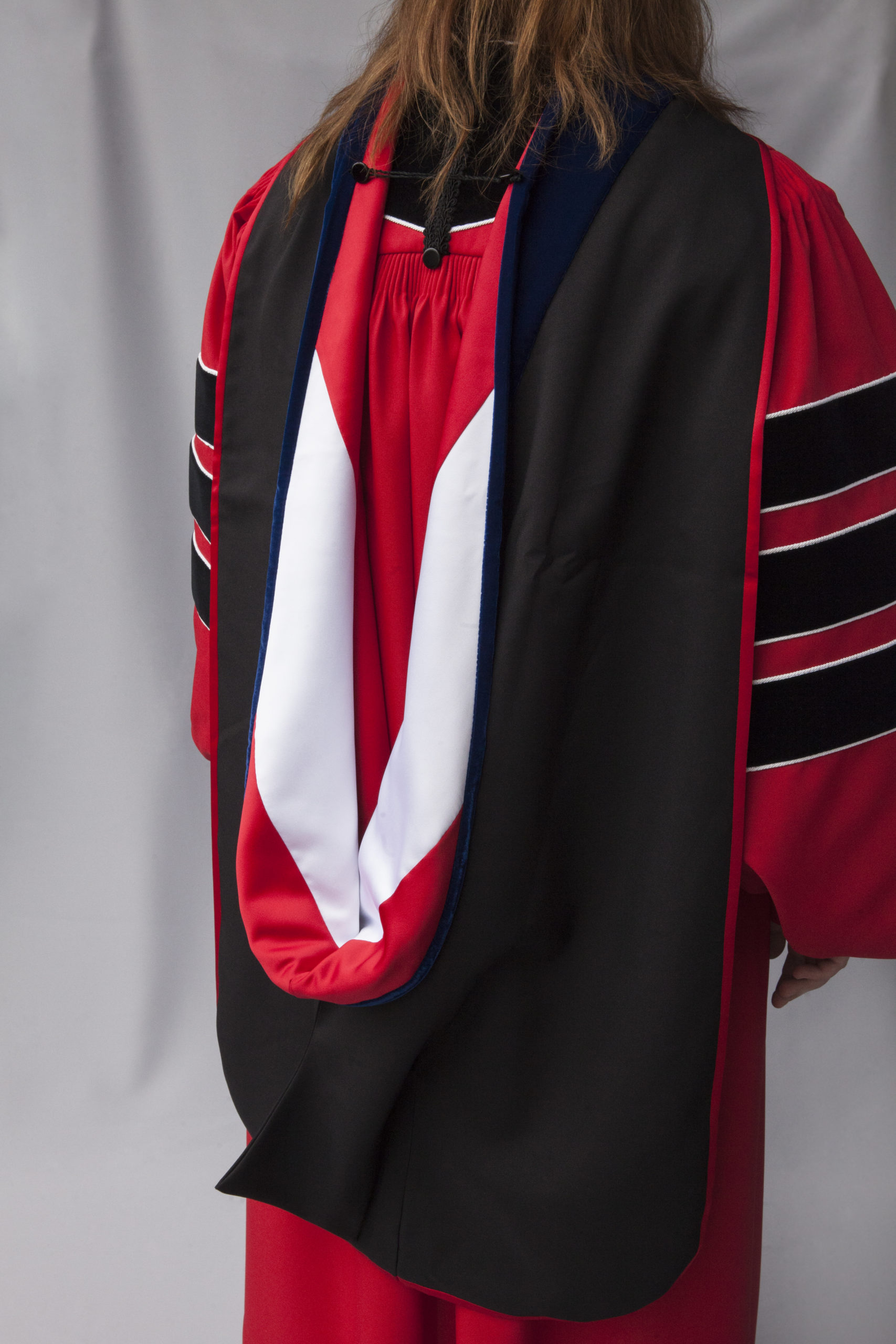 A rectangular, black velvet hood with a red field and white chevron worn by a person also wearing a Phd Red Robe with black bars on the sleeves.